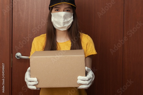 Coronavirus protection. Safe Delivery of orders from online stores. Young woman courier in uniform in a medical antibacterial mask and gloves stands at the door with a box photo