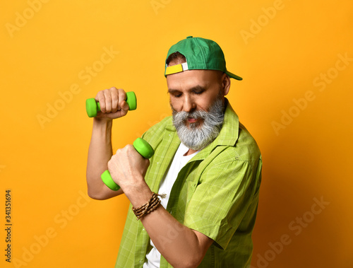 Aged man in green cap and shirt, white t-shirt, bracelet. He doing exercises with two dumbbells, posing on orange background