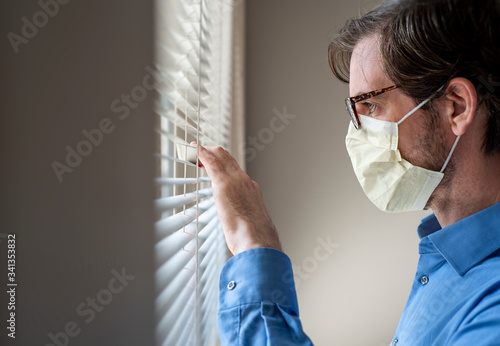 Side profile view of man wearing respirator face mask looking out window through blinds