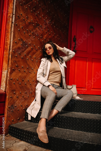 
Street fashion portrait of a young elegant luxurious lady in a beige trench coat, fashion sunglasses, shawl, posing on the street of a European city, against the background of a red building