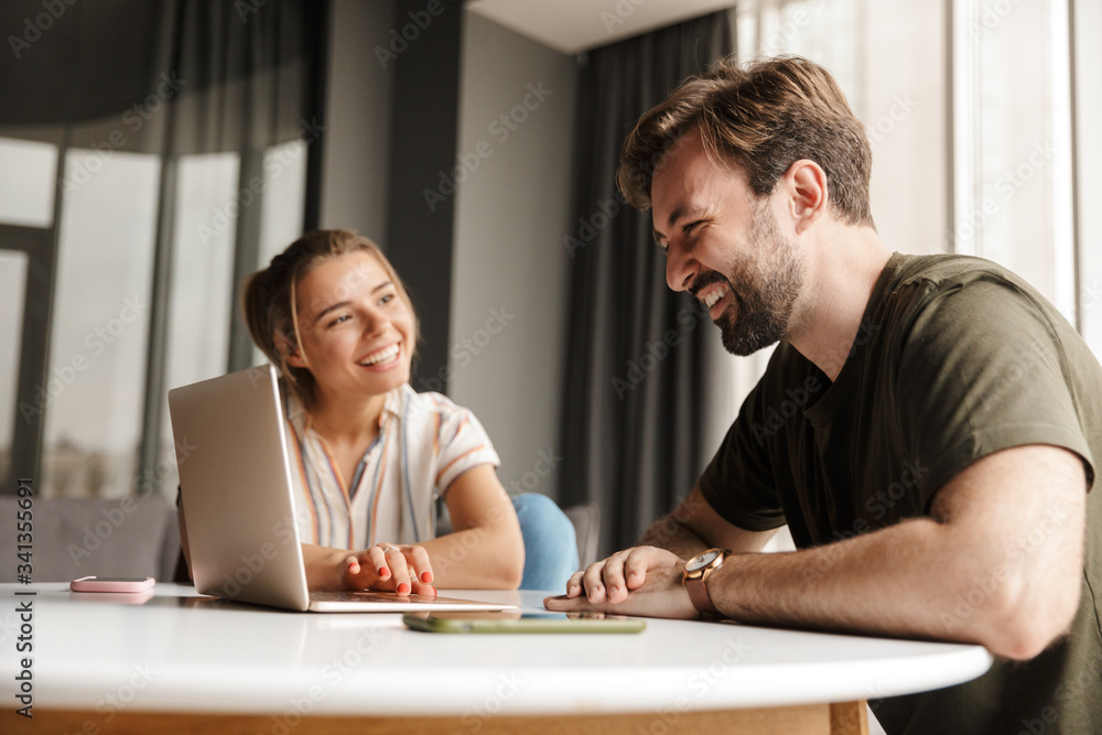 Photo of young joyful couple using laptop and smiling while sitting Photo of young joyful couple using laptop and smiling while sitting