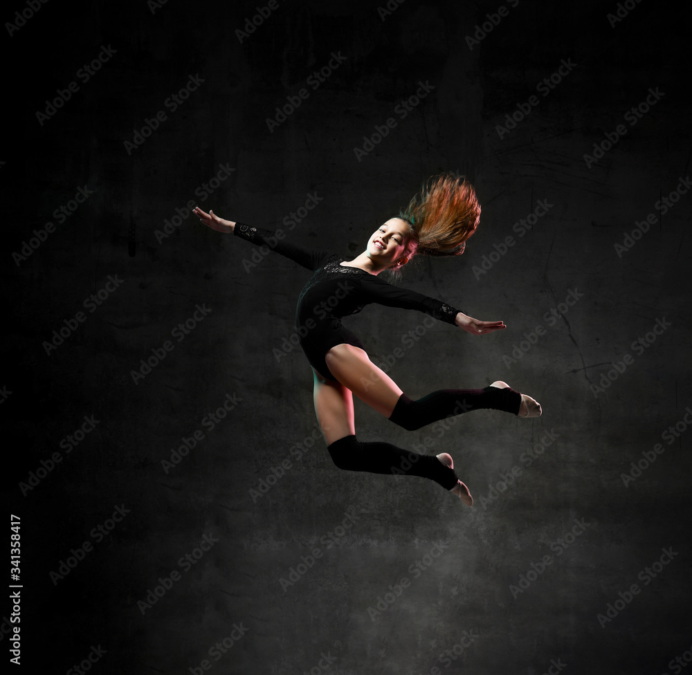 girl gymnast in black sport body and uppers jumping and making dymnastic pose in air over dark background