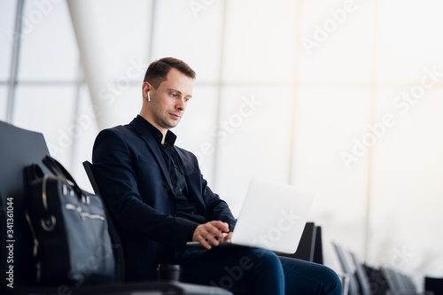 Man with suitcase sitting in airport waiting area while listening music using airpods
