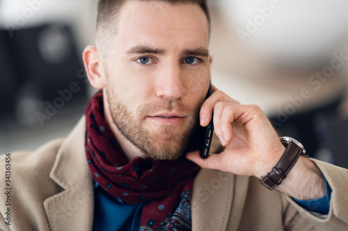 Attractive young adult man in stylish scarf and coat sitting inside airport terminal waiting area. He looking at cellular mobile phone screen and smiling
