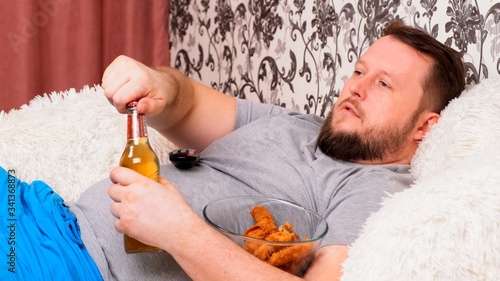 bearded Fat man with a big belly lies on the sofa with fast food, opens a beer and misses the TV with the remote control in his hand, close-up.