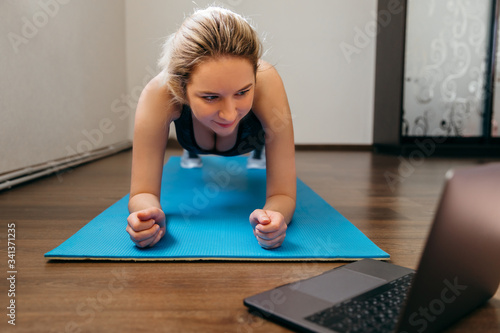 Girl training at home, doing plank and watching videos on laptop, training in living room