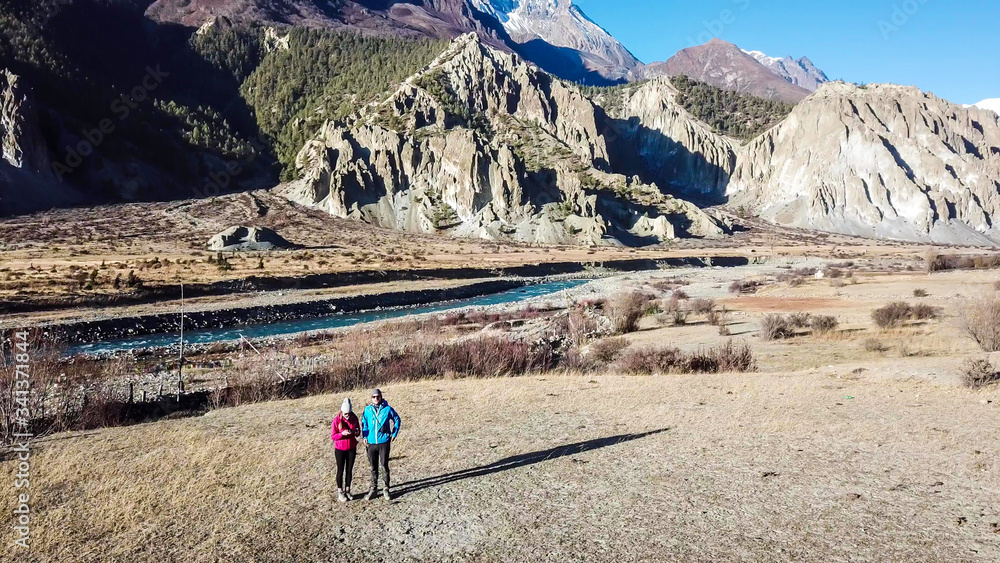 Obraz premium A couple walking on gravelled road, along the Annapurna Circuit Trek, Himalayas, Nepal. Snow caped Annapurna chain in the back. Clear weather, dry grass, snowy peaks. High altitude, massive mountains.