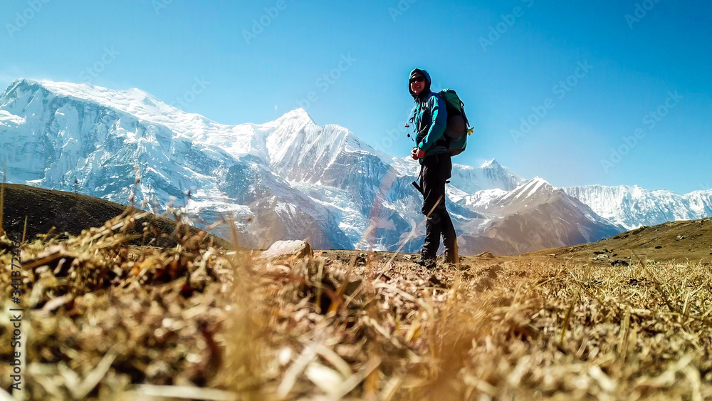 A man trekking on the Annapurna Circuit Trek, Himalayas, Nepal. Snow caped Annapurna chain in the back. Clear weather, dry grass, snowy peaks. High altitude, massive mountains. Freedom and adventure