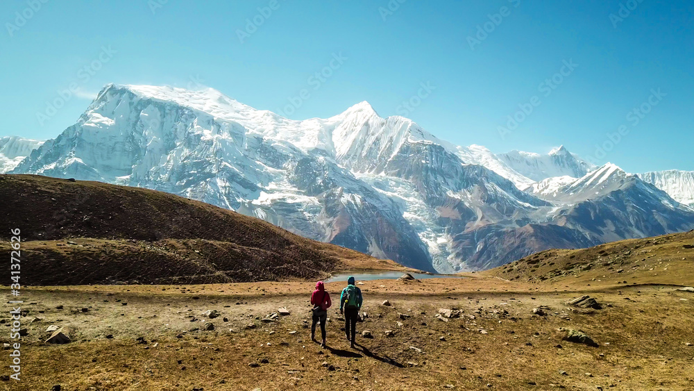 A couple walking on the Annapurna Circuit Trek, Himalayas, Nepal. Snow caped Annapurna chain in the back. Clear weather, dry grass, snowy peaks. High altitude, massive mountains. Freedom and adventure