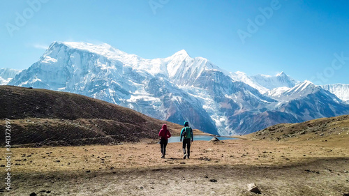 A couple walking towards the Ice lake, along Annapurna Circuit Trek, Himalayas, Nepal. Snow caped Annapurna chain in the back. Clear weather, dry grass, snowy peaks. High altitude trekking. Freedom