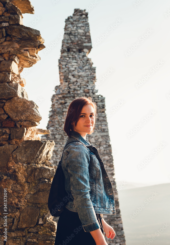 Portrait of young and beautiful brunette girl (woman) with pretty smile in the ancient ruins on sunset. Vertical photo of model with big dark eyes and blue jean jacket turning and posing to camera.