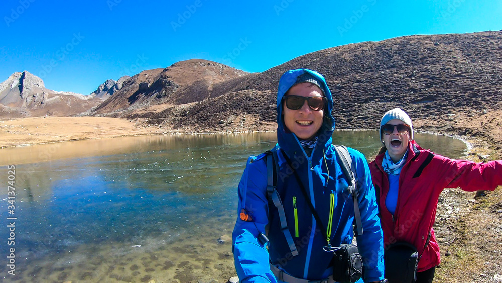 A couple trekking along the Ice Lake, Annapurna Circuit Trek detour, Himalayas, Nepal, surrounded by high mountains. High mountain peaks around them. High altitude lake. Harsh landscape.