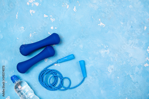 Blue dumbbells, jump rope, bottle of water on blue background. Sport, training, slimming, healthy lifestyle, fitness, home workout concept. Sport equipment. Copy space