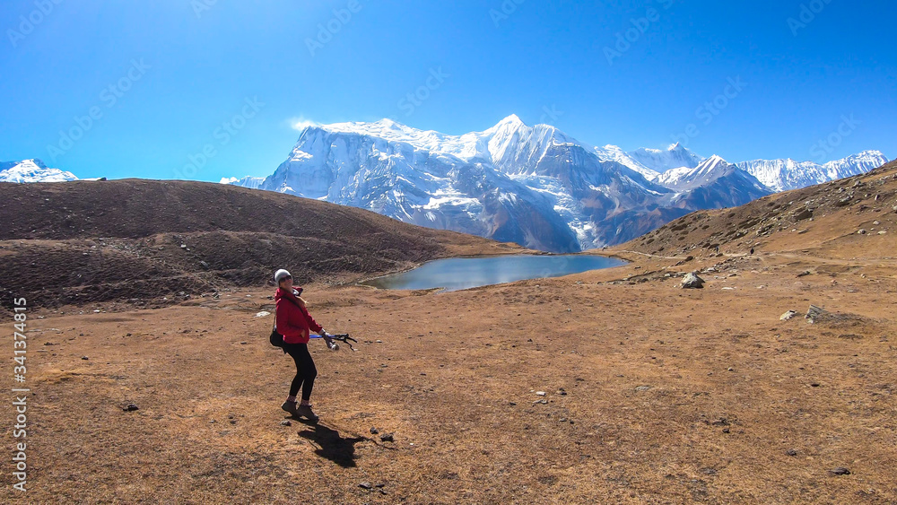 A woman trekking towards Ice Lake, Annapurna Circuit Trek in Himalayas, Nepal, surrounded by high, snow caped Annapurna Chain in the back. High altitude lake. Harsh landscape. She is enjoying the view