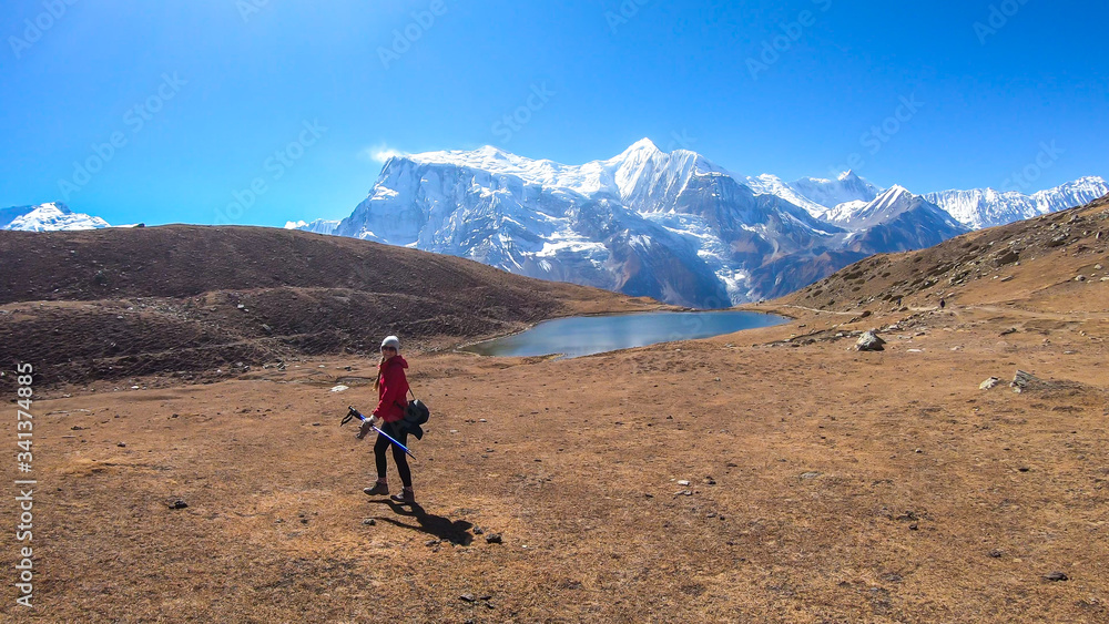 A woman trekking towards Ice Lake, Annapurna Circuit Trek in Himalayas, Nepal, surrounded by high, snow caped Annapurna Chain in the back. High altitude lake. Harsh landscape. She is enjoying the view