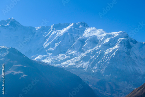 A close up view on snow caped Himalayan peak seen from Annapurna Circuit Trek, Nepal. Sharp and steep slopes of the mountain. Powder snow being blown by strong wind. First sunbeams reaching the peak