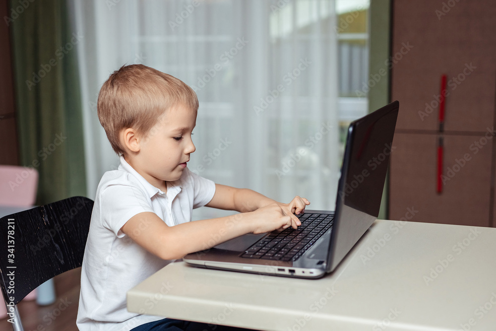 home-based distance education of children during quarantine. happy baby boy in white polo doing homework using laptop sitting at home in the kitchen home-based distance education of children during quarantine. happy baby boy in white polo doing homework using laptop sitting at home in the kitchen