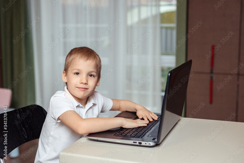 home-based distance education of children during quarantine. happy baby boy in white polo doing homework using laptop sitting at home in the kitchen home-based distance education of children during quarantine. happy baby boy in white polo doing homework using laptop sitting at home in the kitchen