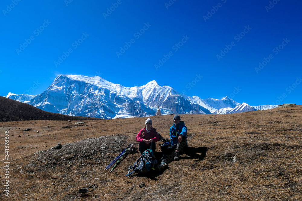 Fototapeta premium A couple sitting on the ground along Annapurna Circuit Trek, Himalayas, Nepal. They are having a trekking break. High mountains covered with snow behind them. The grass is dried. High altitude.