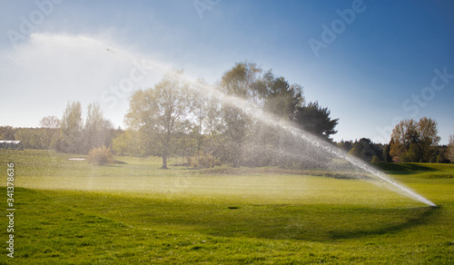 A Sprinkler fountain shooting water over a golf course with green grass and a blue sky.  © Ben