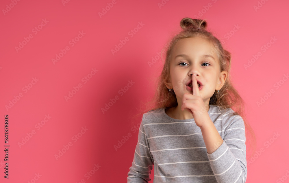Little blonde child with bun hairstyle, dressed in gray striped blouse. She showing be quiet sign, posing against pink studio background. Childhood, fashion, advertising. Close up, copy space Little blonde child with bun hairstyle, dressed in gray striped blouse. She showing be quiet sign, posing against pink studio background. Childhood, fashion, advertising. Close up, copy space