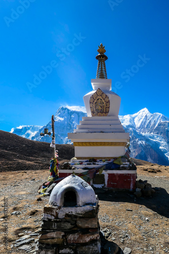 A stupa with Annapurna Chain as a backdrop, Annapurna Circuit Trek, Himalayas, Nepal. High mountains covered with snow. Land in front of the stupa is barren and dry. Some prayer's flag next to it.