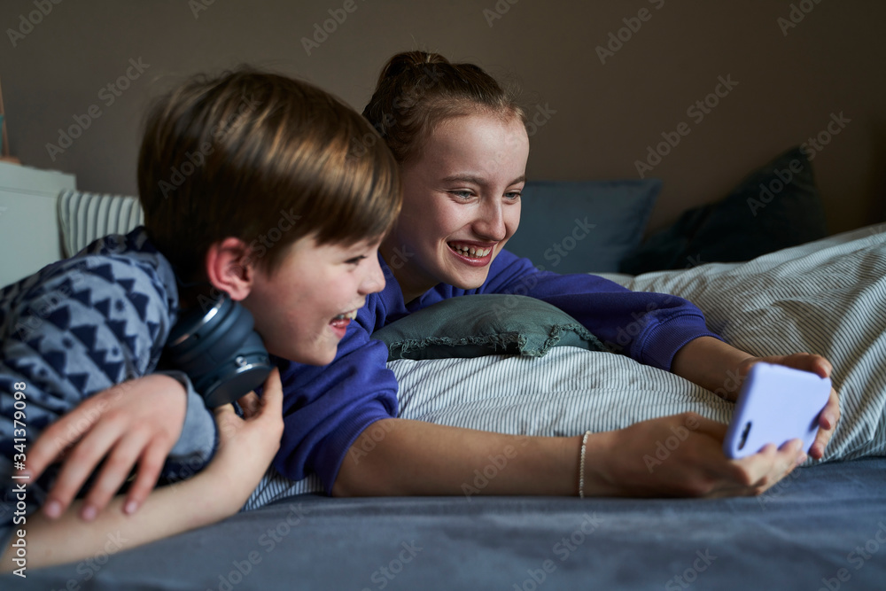 Laughing sibblings lying together on bed using smartphone for video chat Laughing sibblings lying together on bed using smartphone for video chat