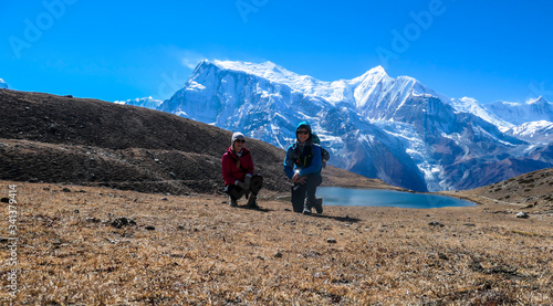 A couple in hiking outfits squatting next to a huge rock next to Ice Lake, Annapurna Circuit Trek, Himalayas, Nepal. High, snow caped Annapurna chain in the back. Happiness and love
