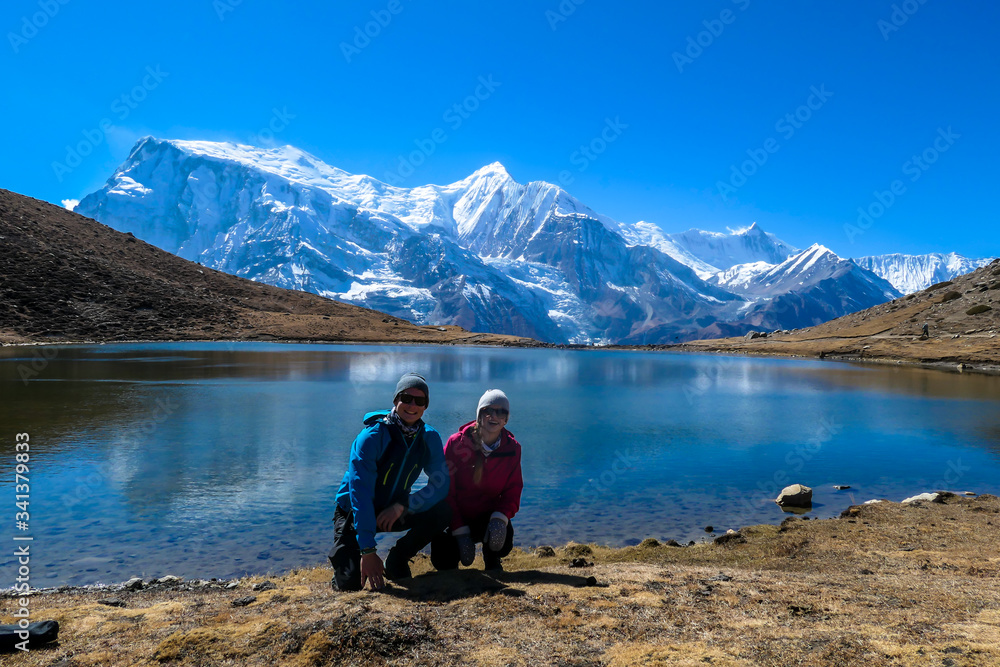 A couple in hiking outfits squatting at the shore of Ice Lake, Annapurna Circuit Trek, Himalayas, Nepal. They are having fun. High, snow caped Annapurna chain in the back. Happiness and love