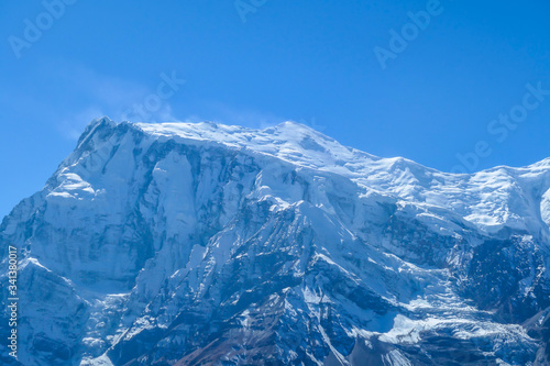 A close up view on snow caped Himalayan peak seen from Annapurna Circuit Trek, Nepal. Sharp and steep slopes of the mountain. Powder snow being blown by strong wind. First sunbeams reaching the peak