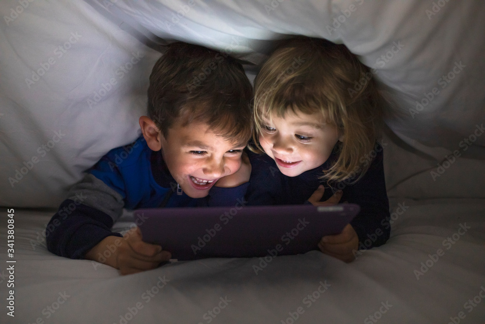 Portrait of brother and his little sister lying side by side under a blanket having fun with digital tablet Portrait of brother and his little sister lying side by side under a blanket having fun with digital tablet