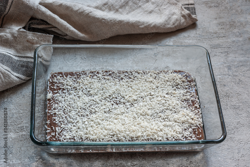 Brown (chocolate) dough in a rectangular glass baking dish sprinkled with coconut. On a gray concrete table next to a beige towel. Cooking process Brown (chocolate) dough in a rectangular glass baking dish sprinkled with coconut. On a gray concrete table next to a beige towel. Cooking process