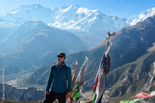 A man in hiking outfit standing on a barren pathway along Annapurna Circuit Trek. There are few prayer flags next to him. Snow caped Annapuna chain in the back. Achievement and completion