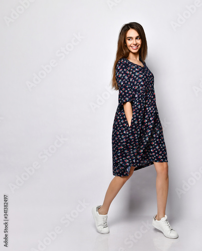 Brunette model in black dress with floral print, white sneakers. Smiling, touching her face, posing isolated on white. Full length © FAB.1