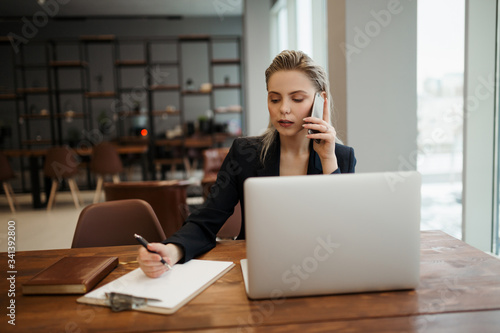 A young girl-Manager, sitting in a coworking office at her workplace in front of a laptop, and talking intently on the phone