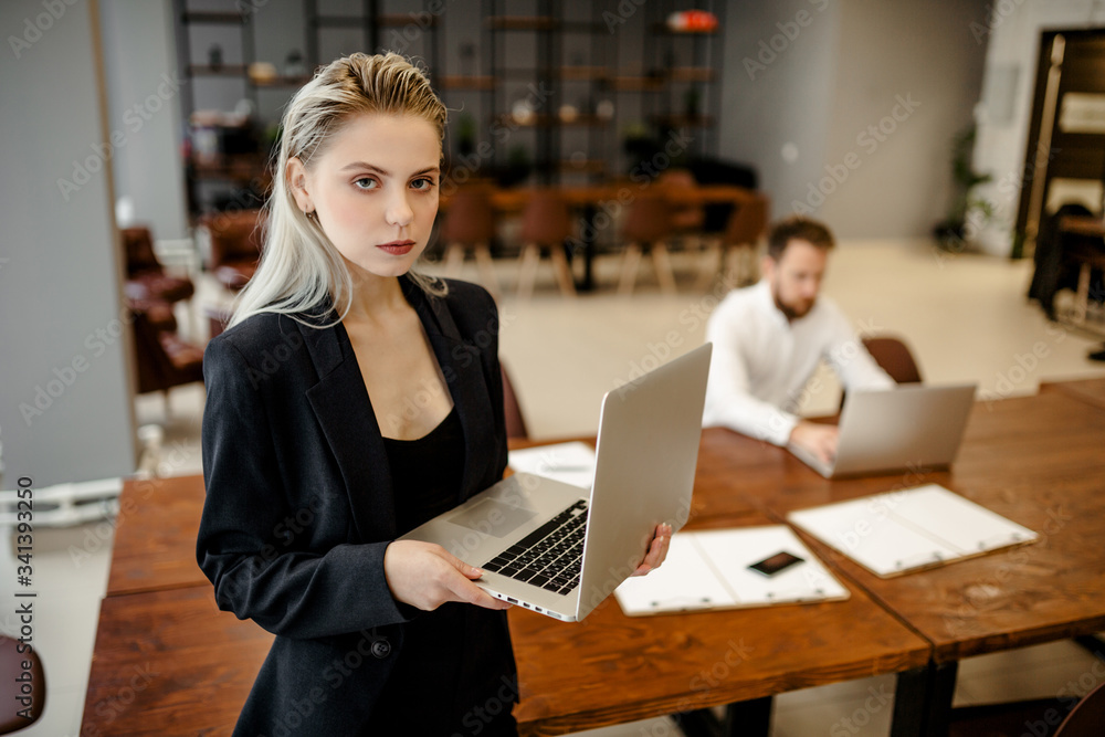 A young Manager girl standing in the office next to the workplace, with a laptop in her hands and looking directly at the camera A young Manager girl standing in the office next to the workplace, with a laptop in her hands and looking directly at the camera