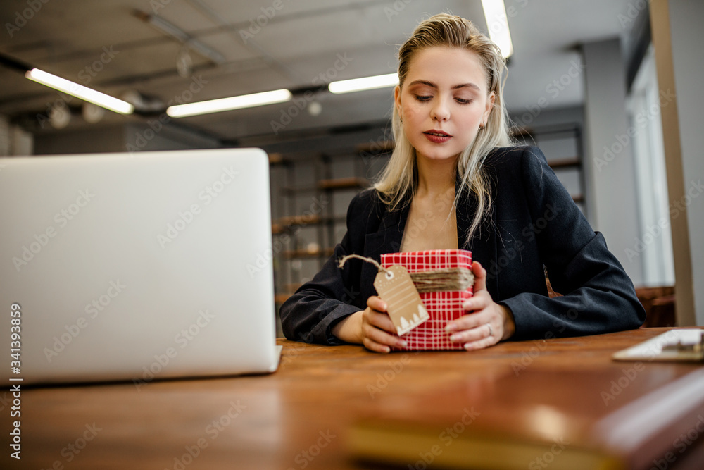A beautiful blonde businesswoman sits at a Desk in the office and holds a gift that was given by colleagues at work A beautiful blonde businesswoman sits at a Desk in the office and holds a gift that was given by colleagues at work