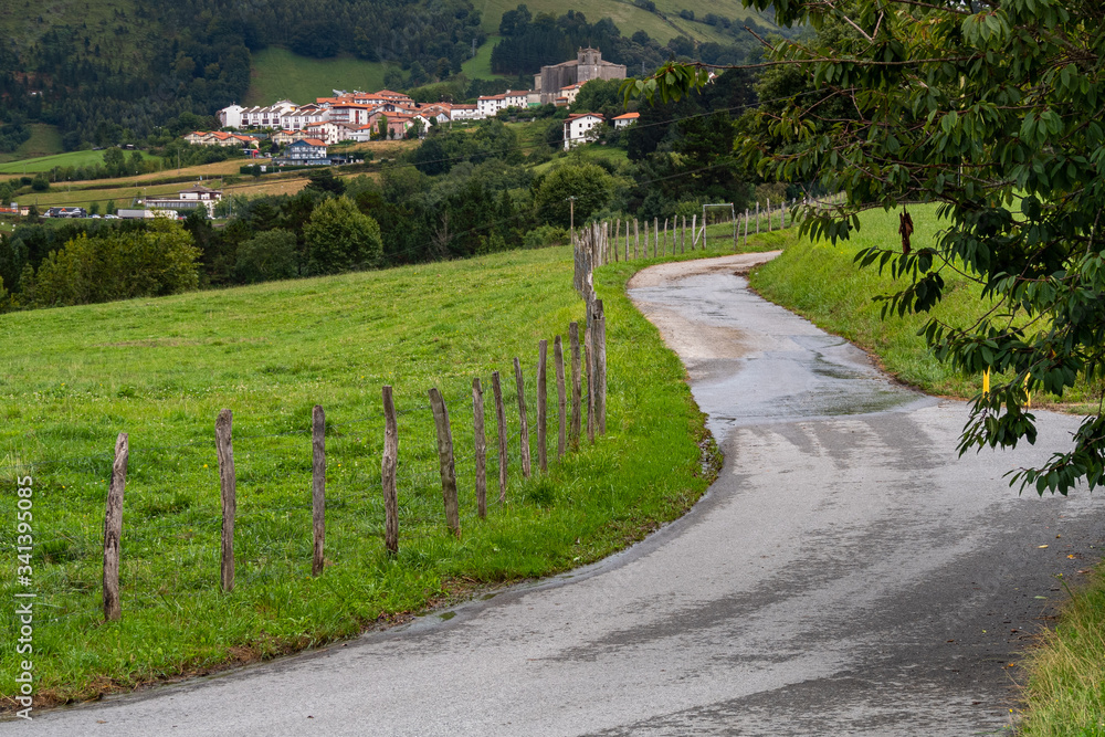 Winding rural country road goes along fence with typical rural village and church of Basque Country, Spain. Camino de Santiago. Hiking and traveling concept.