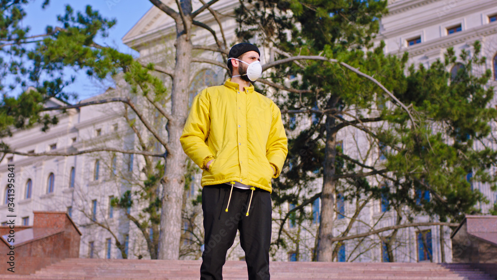 Young man in a yellow jacket and hat wearing his protective mask while standing on the park stairs new Covid-19