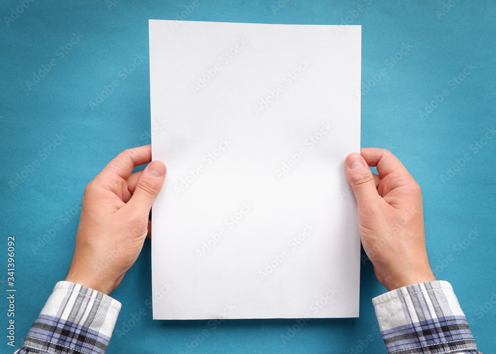 Hands of a man in a plaid shirt holding a sheet of white blank paper on a blue background. Hands of a man in a plaid shirt holding a sheet of white blank paper on a blue background.
