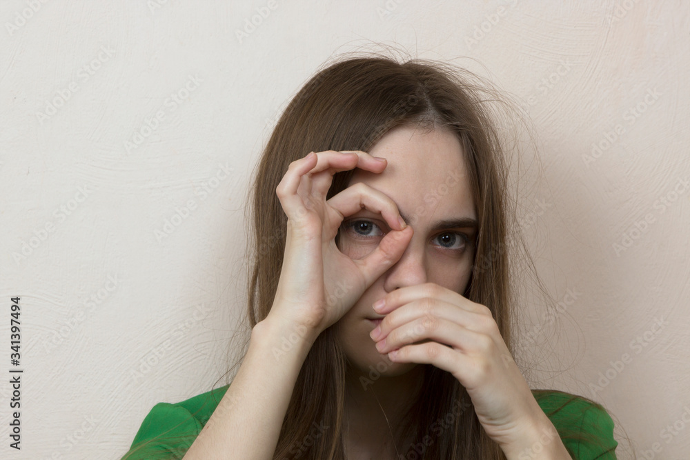 a girl with long blond hair covers her mouth with one hand, and with a second hand shows an ok sign. Teenager misses self-isolation in an apartment. a girl with long blond hair covers her mouth with one hand, and with a second hand shows an ok sign. Teenager misses self-isolation in an apartment.