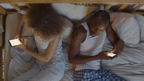 Flat lay of African man wearing pajamas and his wife with curly hair lying back to back in bed and using their mobile phones