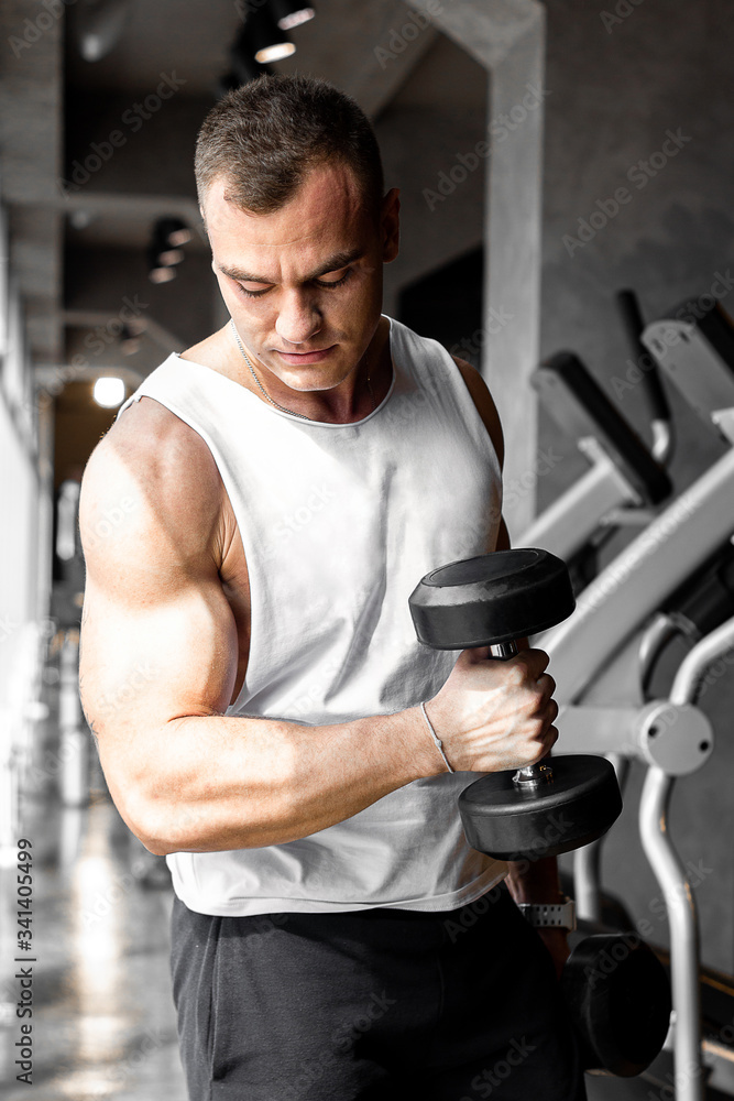 Bodybuilder doing biceps exersices with dumbbell in gym around fitness equipment and sunrays laying on his muscule arm. Bodybuilder doing biceps exersices with dumbbell in gym around fitness equipment and sunrays laying on his muscule arm.