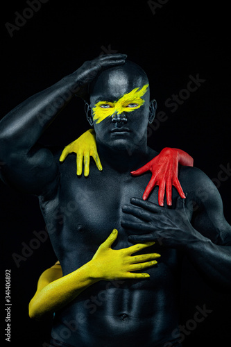 Fototapeta Naklejka Na Ścianę i Meble -  Man with female hands on the body. Bodybuilder athlete with yellow face art and body paint. Colorful portrait of the guy with bodyart.