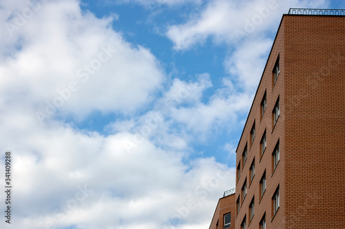 The wall of a high-rise building made of brick against the blue sky in the clouds. Corner view of a brick building against a blue sky. Windows on the facade of a high-rise building, side view.