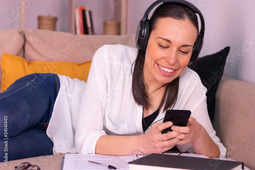 Pretty brunette young woman smiling with her smartphone studying stretched out on the sofa at home listening to music through headphones. She is wearing a white shirt and blue jeans.