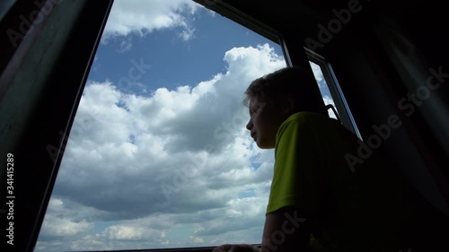 Silhouette of young white kid watching pensively out through glass of closed window standing in room indoors. Cloudy sunny blue sky moving outdoors. Timelapse video. photo