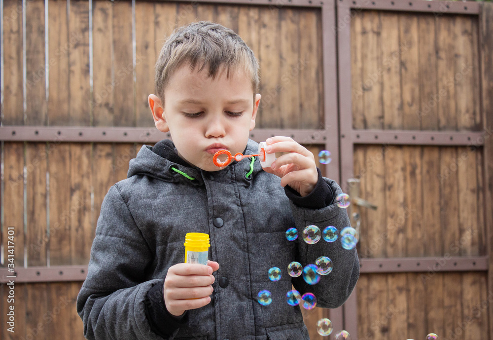 Funny brown-haired child boy in a gray jacket blowing soap bubbles from a special ring on a wooden background. Soap foam in yellow packaging. Good family vacation Funny brown-haired child boy in a gray jacket blowing soap bubbles from a special ring on a wooden background. Soap foam in yellow packaging. Good family vacation