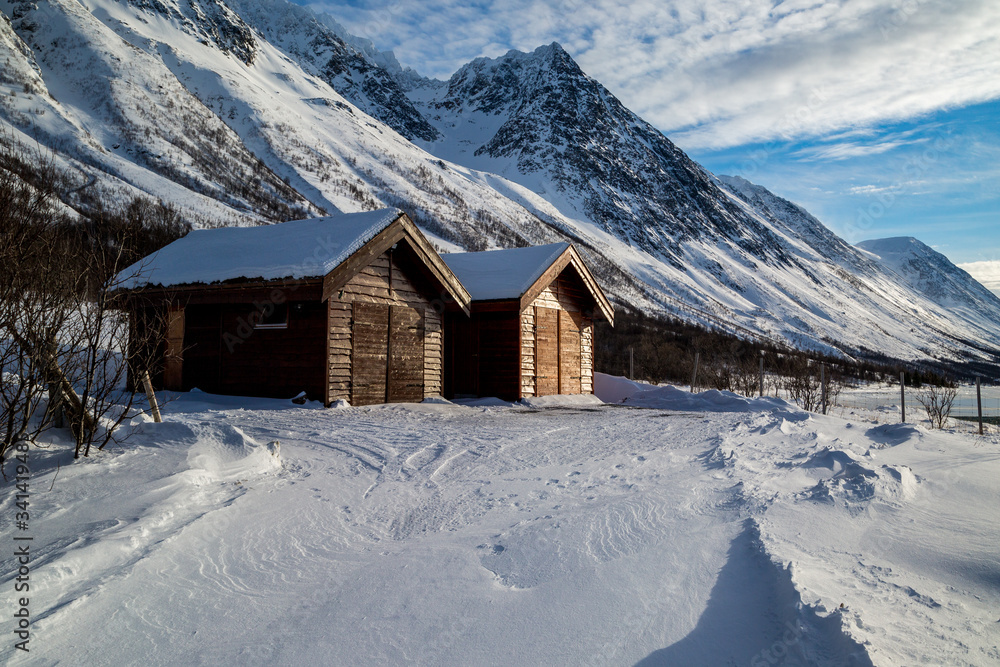 Wooden sheds along a road covered by snow drifts glowing in the afternoon sun on the shore of a fjord in the Lyngen Alps in Northern Norway.
