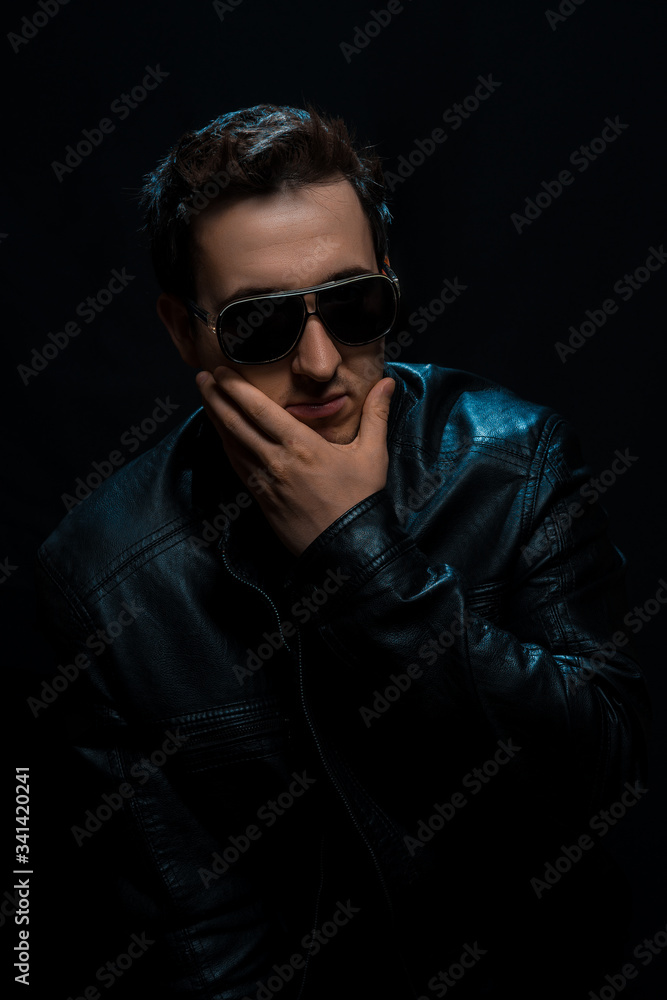 Obraz premium Young sexy stylish guy with glasses in a black leather jacket on a black background